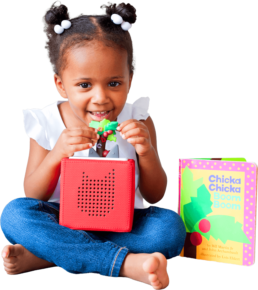  A young girl with her hair in buns sits cross-legged on the floor, smiling as she places a 'Chicka Chicka Boom Boom' coconut tree figurine onto the top of a red Toniebox. To her right sits a matching 'Chicka Chicka Boom Boom' board book.