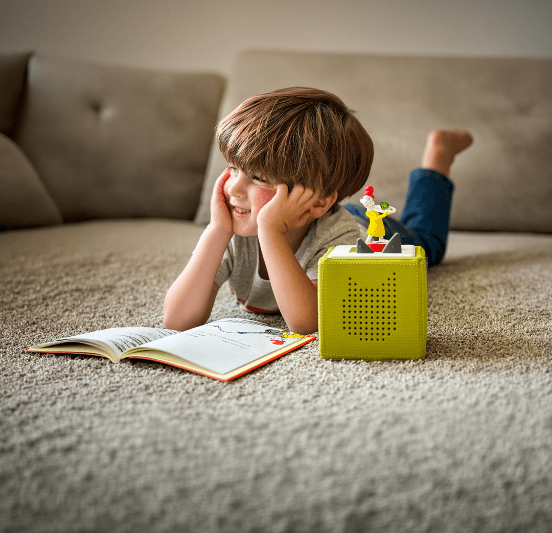 A young boy lies on his stomach on a plush carpet, propping his chin in his hands with a joyful expression. In front of him is an open Dr. Seuss book and a lime green Toniebox with a Sam-I-Am figurine placed on top, creating an interactive reading and listening environment.