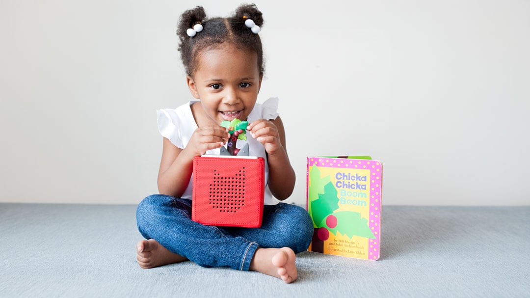 Young girl holding a red Toniebox audio player next to the book Chicka Chicka Boom Boom by Bill Martin Jr and John Archambault.
